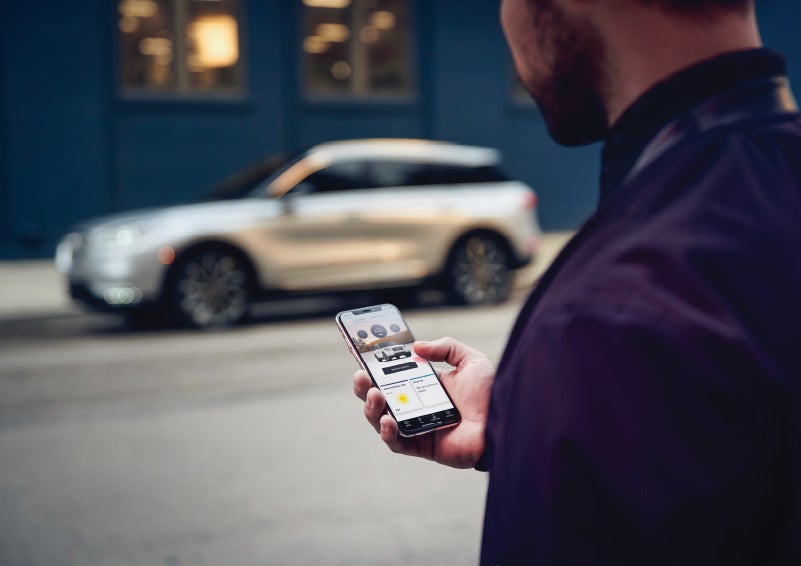 A man is holding his phone looking at the Lincoln Way™ App and we see his 2022 Lincoln Corsair SUV in the background against a blue brick wall | Mark Ficken Lincoln in Charlotte NC