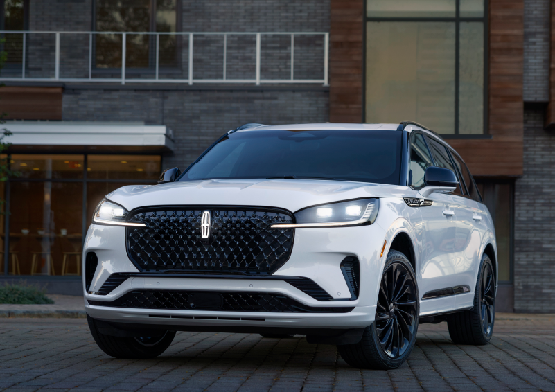 A white 2025 Lincoln Aviator® SUV is shown parked outside a modern building with gray brick and wood accents. | Mark Ficken Lincoln in Charlotte NC